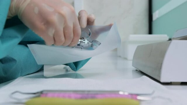 Woman doctor assistant in white gloves and green gown puts dental tools into craft package for further disinfection in autoclave. Side view of dentist prepares instrument syringe for sterilization