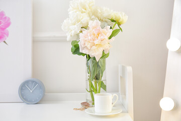 Vase with beautiful peony flowers, cup of coffee and alarm clock on table in room