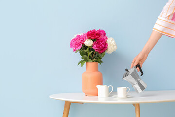 Woman pouring hot coffee from pot into cup on table with beautiful peony flowers