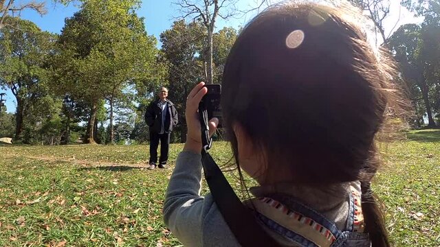 Asian little child girl photographer takes pictures of her grandfather in a summer park. Concepts of technology and hobby lifestyle for child.