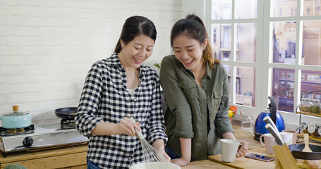 two beautiful asian japanese ladies friends baking cake in home kitchen. charming women laughing while preparing dough in bowl with whisk. roommate stand beside looking and talking with cup of tea.