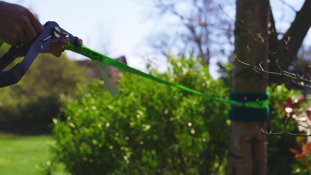Over the shoulder of a man tightening bright green slack line outside near green bush on lush grass with trees and barn in the background. Blue sky with some cloud.