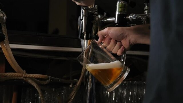 The bearded bartender with a tattoo filling a glass with beer while standing at bar counter in pub. Barman hand at beer tap pouring a draught lager beer.