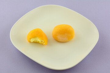 Two mango cream mochi balls one with missing bite on white plate on lavender background