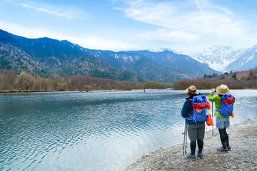 Fototapeta premium Hikers wearing colourful attire stop and photograph Lake Taisho pond and the distant snowy mountain peaks of Chubu Sangaku National Park in Kamikochi, Nagano Prefecture, Japan. Selective focus.