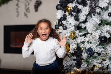 Beautiful little girl standing at the large fireplace and holding a gift, in the bright New Year's interior
