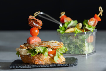 a tasty looking open sandwich with cheese, assorted meat and vegetables with an out of focus stainless steel serving basket with vegetables