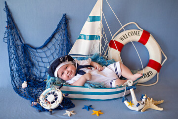 Little baby boy lies in a boat in sailor clothes. A child in the form of a sailor. Lifebuoy, fishing net with shells.