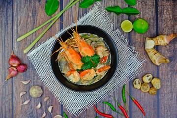 Authentic Tom Yum Kung, popular and delicious Thai hot spicy soup with shrimp, mushrooms and Thai herbs condiments on wooden table surrounded by key ingredients.