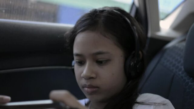 Cute asian young girl wearing headphones and using mobile phones during traveling in a car. Female teenager watching online movies while sitting in the back seat of a car. 