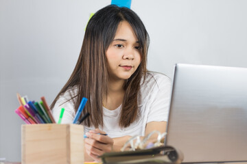 Asian women wear a white T-shirt. Working at home is comfortable And eyes looking at the computer screen in a grey laptop Resting on the desk And the pen handle and the white wall background