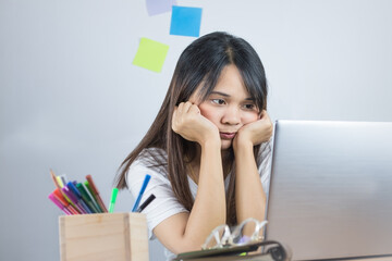Asian women wear white shirts. Working at home feeling stressed and eyes looking at the grey laptop computer screen on the table and hands holding the chin. And white wall background