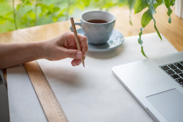 Hand writing something on piece of paper, ruler, laptop, cup of tea on the desk