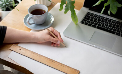 Hand writing something on piece of paper, ruler, laptop, cup of tea on the desk