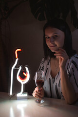 Beautiful girl with a glass of wine on the background of a neon sign of a wine bottle and a glass. Trendy stylish. Glowing life. Modern style. Neon sign.