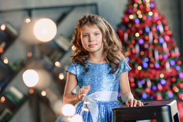 girl with dark hair standing on a box with gifts. Christmas tree in the background. smiles