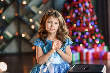 girl with dark hair standing on a box with gifts. Christmas tree in the background. smiles