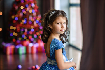 girl with dark hair standing on a box with gifts. Christmas tree in the background. smiles