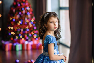 girl with dark hair standing on a box with gifts. Christmas tree in the background. smiles