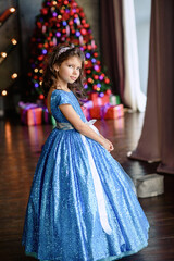 girl with dark hair standing on a box with gifts. Christmas tree in the background. smiles