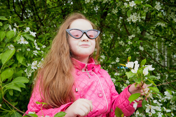 Pretty cute little girl with long hair and in pink glasses posing near a blooming apple tree with white flowers in a summer park