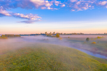Obraz premium Morning fog in the meadows and at the mouth of the river during sunrise in the countryside, view from the height of the landscape on the horizon forest and village.