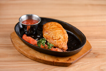 Juicy grilled chicken fillet with tomato sauce served in a black tray over rustic wooden background.