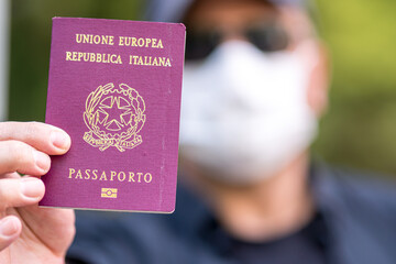 Travelling during the coronavirus disease outbreak. Tourist wearing a medical protective mask showing to the camera his Italian passport. Selective focus