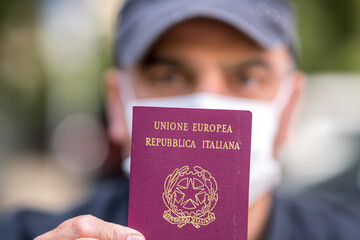 Travelling during the coronavirus disease outbreak. Tourist wearing a medical protective mask showing to the camera his Italian passport. Selective focus
