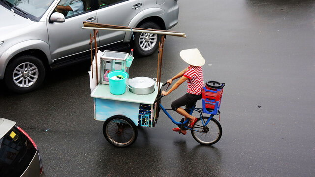 Vietnamese woman ride three wheel bicycle on wet street, transfer mobile restaurant of food vendor