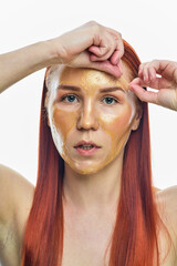 a close portrait of a red-haired girl who removes a gold cosmetic mask from her face. Isolated on a white background.