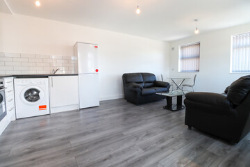 A modern British apartment living room and kitchen with black leather sofa and a coffee table on a grey wooden floor with white walls and modern fitted kitchen with a fridge