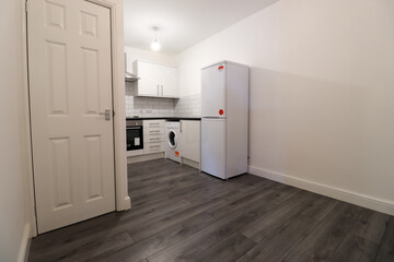 A modern British apartment kitchen with a grey wooden floor with white walls and modern fitted kitchen with washing machine, cooker stove and white fridge.