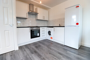 Fototapeta premium A modern British apartment living room and kitchen with black leather sofa and a coffee table on a grey wooden floor with white walls and modern fitted kitchen with a fridge