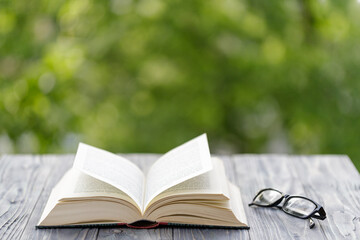 Open book and glasses on the wooden table. Blurred green tree background with copy space