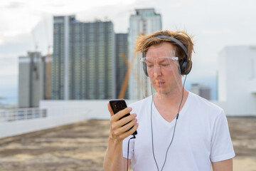 Fototapeta na wymiar Young handsome man with face shield using phone and listening to music at rooftop of the building