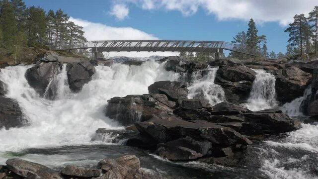 Likholefossen waterfall in Norway. A turbulent mass of raging water falling from the rocks, foaming, and splashing. A narrow metal bridge standing above the river. Water haze rising above the surface.