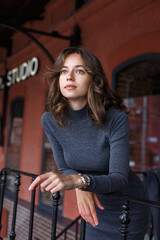 Fototapeta na wymiar young thoughtful pretty girl in gray turtleneck, skirt with watches on arm poses in front of red building. Street style portrait photosession of attractive female, urban photoshoot of elegant model