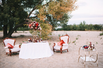 A banquet on nature, a table with a white tablecloth, vintage plates with a gold rim, wine glasses, cutlery, candles, a composition of flowers, amaryllis, roses, red dahlias.
