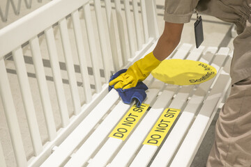 Housekeeper cleaning public chair with social distancing sign on chair by towel and gloves. COVID-19 prevention sanitizing inside.