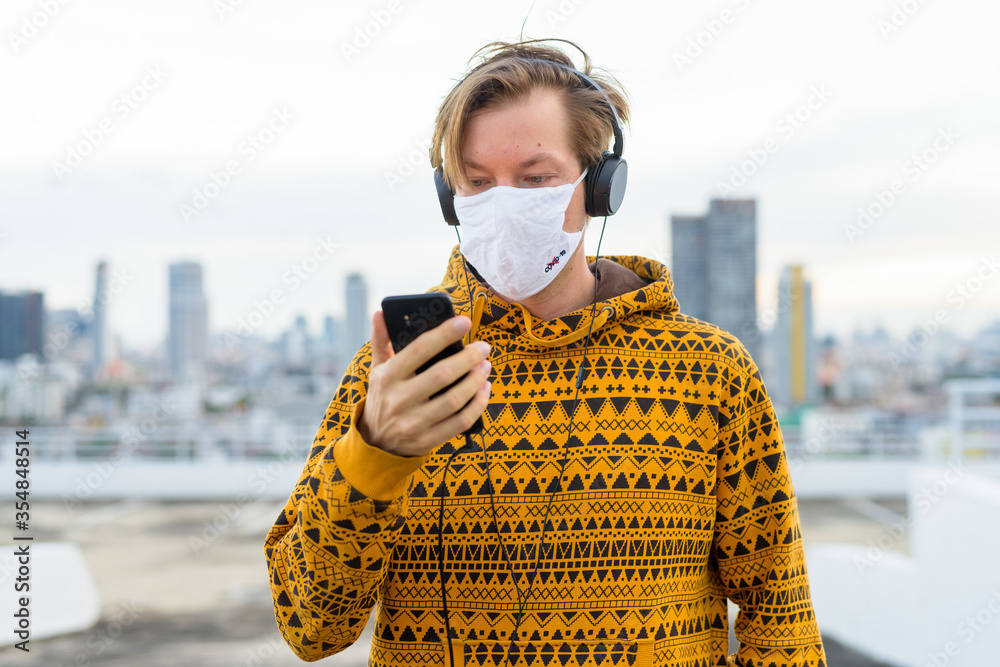 Wall mural Young man in hoodie using phone while listening to music with mask against view of the city