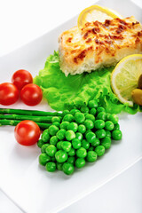 Cod fillet baked with cheese and onions with a garnish from an asparagus and green peas. On white background. High key. Selective focus.