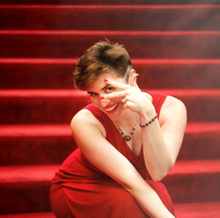 Emotional pretty stylish short-haired woman in red dress sitting on red stairs. Happy elegant playful girl with short haircut sit on footsteps. Luxury concept. Makeup and jewellery on beautiful lady.