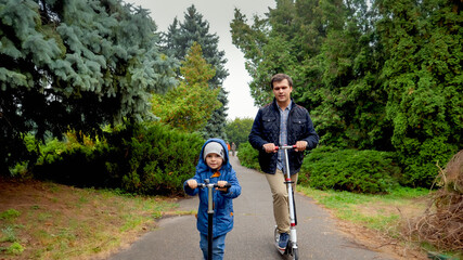 Happy smiling toddler boy riding with young father on kick scooter at autumn park