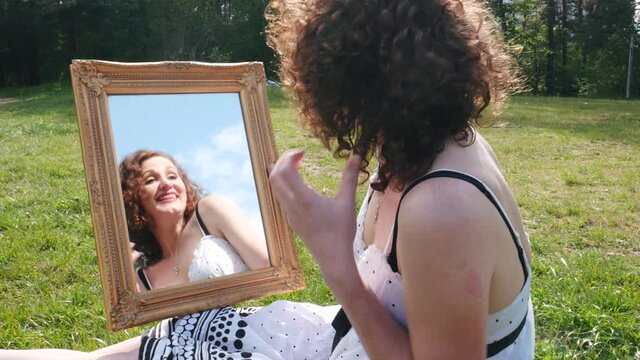 Funny charismatic artistic beautiful female actress in white dress looking at mirror and enjoying herself, sitting outdoors on a green field in summer