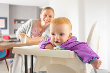Happy red haired baby and her mom having fun during family dinner. Little child sitting in high chair spotted with food. Feeding or child care at home concept