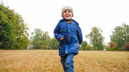 Portrait of laughing little boy running on grass at field on cloudy autumn day