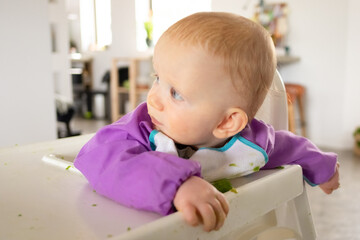 Adorable baby sitting in high chair with puree smudges, looking away. Closeup shot. First solid food or child care at home concept
