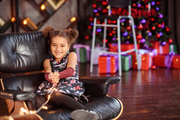 Moscow, Russia 2018 01 December  Beautiful kid girl 5-6 year old wearing stylish dress sitting in armchair over Christmas tree in room. Looking at camera. Holiday season.