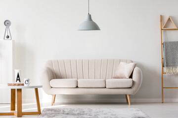 Grey lamp above pillow on white sofa next to wooden coffee table with mugs, copy space on empty wall